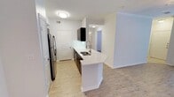 This single-floor space showcases a contemporary open-concept layout that seamlessly connects the kitchen to the living area. The kitchen is a standout feature, anchored by a white quartz peninsula that doubles as a breakfast bar and prep space, complete with an integrated sink and a sleek gooseneck faucet. Dark cabinetry provides a striking contrast against the bright white countertops and walls, while upper cabinets in a deep finish add depth to the design. Stainless steel appliances, including a French door refrigerator, contribute a polished, modern aesthetic. Light wood-look flooring runs throughout the space, adding warmth while remaining easy to maintain. Recessed flush-mount ceiling fixtures provide consistent overhead lighting, and crown molding along the upper walls introduces a refined architectural detail. A hallway visible from the kitchen leads to additional rooms, hinting at a thoughtful floor plan. The overall palette of crisp whites, dark accents, and natural wood tones creates a clean, contemporary ambiance suited for everyday living.