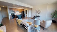 Located in Liberty Lake, Washington, this single-floor residence offers a thoughtfully designed open-concept layout that seamlessly connects the living, dining, and kitchen areas. The living room is bright and welcoming, with large windows that flood the space with natural light and provide views of the surrounding neighborhood. Track lighting overhead adds a modern touch, complementing the light wood-style flooring that extends throughout the main living areas.

The kitchen is a standout feature, outfitted with warm wood cabinetry, light countertops, a double-basin sink, and black appliances including a gas range and dishwasher. A freestanding island with a rich wood top and built-in wine storage offers additional prep space and casual seating. A dedicated laundry area with a washer is conveniently tucked nearby.

The bedroom is carpeted for comfort and features a window that brings in natural light. The bathroom is designed with accessibility in mind, including sturdy grab bars and a handheld showerhead in a clean, white walk-in shower. A separate room visible in the floor plan offers flexible space, suitable for a home office or den. Throughout the residence, neutral tones and clean lines create a calm, functional ambiance well-suited for everyday living.