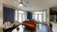 This single-story space showcases an open-concept layout that seamlessly connects the living and dining areas, creating a cohesive and functional environment. Rich, dark hardwood flooring runs throughout, providing a striking contrast against the light-toned walls and bright natural light that floods the space through multiple large windows and glass-paneled doors. Patterned drapes frame the windows, adding a layer of visual interest and privacy.

The living area features a tufted burnt-orange sofa as its centerpiece, complemented by a striped accent chair and a round white coffee table. A ceiling fan with a built-in light fixture is centrally positioned overhead, contributing to both comfort and ambiance. The space carries a distinct mid-century modern aesthetic.

Adjacent to the living area, the dining space accommodates a rectangular table with upholstered chairs, lit by a contemporary track-style chandelier. Additional glass-paneled doors in the dining area allow further natural light and provide access to the exterior. Together, the areas create a warm, inviting atmosphere well-suited for both everyday living and entertaining.