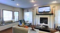 This single-floor space centers around a well-appointed living room that blends comfort with functional design. The room features a built-in fireplace with a sleek dark surround and mantel, above which a wall-mounted flat-screen television is positioned for optimal viewing. The textured ceiling hosts recessed lighting that complements the natural light streaming through multiple windows dressed with white sheer curtains and horizontal blinds, offering both brightness and privacy. The neutral gray wall tones create a versatile backdrop, accented by warm décor elements including decorative branches and floral arrangements. Seating is thoughtfully arranged, with a plush sofa and a teal accent armchair adorned with earthy-toned throw pillows in rust and brown, adding pops of color to the space. A dark wood coffee table serves as a central anchor, styled with a decorative tray and accents. The flooring transitions between carpet and a darker surface near the seating area. The overall ambiance is warm and inviting, balancing casual comfort with curated, stylish touches throughout the living area.