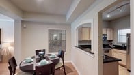This single-floor residence offers a thoughtfully designed layout that balances functionality and comfort. The dining area is a welcoming space, featuring warm hardwood flooring, neutral-toned walls, and a tray ceiling that adds a subtle architectural detail overhead. A window with horizontal blinds allows natural light to filter into the room, creating a bright and inviting atmosphere. The dining space comfortably accommodates a round table with seating, making it well-suited for both everyday meals and casual entertaining. A standout architectural feature is the arched pass-through opening that connects the dining area to the kitchen, adding a classic design touch while maintaining an open, airy feel between the two spaces. The kitchen is visible through the pass-through and showcases light wood cabinetry, dark granite or stone countertops, and stainless steel appliances, offering a well-appointed cooking environment. The cohesive color palette of soft grays and warm neutrals flows seamlessly throughout, giving the home a unified and polished aesthetic. The single-floor layout ensures ease of movement and accessibility across all areas of the space.