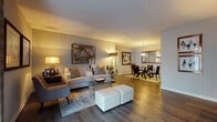 This thoughtfully designed single-floor residence offers a cohesive blend of comfort and style throughout its well-defined living spaces. The open-concept living and dining area serves as the heart of the home, featuring warm wood-look flooring, neutral gray walls, and large sliding glass doors fitted with vertical blinds that allow ample natural light to filter in. The living area is generously sized to accommodate a full seating arrangement, complemented by a glass-top coffee table and upholstered ottomans. The adjacent dining space features a round glass table beneath a statement pendant light fixture, with a large framed mirror adding depth to the room. The kitchen is efficiently laid out with dark cabinetry, stone-look countertops, a mosaic tile backsplash, and black appliances including a full-size refrigerator and dishwasher, with wood-look flooring continuing the cohesive design. The bedroom is a serene retreat with plush carpeting, soft neutral tones, a large window with patterned drapes, and warm ambient lighting. Two full bathrooms are finished with dark vanity cabinetry, stone countertops, and soaking tub-shower combinations. Throughout the space, a consistent warm, neutral palette creates an inviting and polished atmosphere.