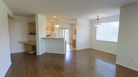 This single-floor space features an open-concept design that connects the living, dining, and kitchen areas into one cohesive, flowing layout. Rich, dark hardwood flooring spans the main living and dining zones, adding warmth and a polished aesthetic throughout. A large window in the dining area fills the space with abundant natural light, complemented by a stylish pendant light fixture overhead that adds a decorative focal point.

The kitchen is positioned centrally and defined by a raised breakfast bar peninsula, offering both a functional workspace and a casual seating area. Light wood cabinetry lines the kitchen walls, providing ample storage, while white appliances including a refrigerator and microwave maintain a clean, cohesive look. A ceiling fan with a light fixture keeps the kitchen area well-ventilated and bright. Sliding glass doors at the rear of the kitchen area allow additional natural light to pour in and suggest access to an outdoor space. The white walls throughout contribute to an airy, neutral palette, making the overall ambiance feel open, clean, and move-in ready.