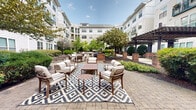 Located at 1901 S Charles St in Baltimore's South Baltimore neighborhood, this beautifully designed outdoor courtyard serves as a central communal gathering space. The grounds feature an expansive interlocking brick paver patio that provides a durable and visually appealing surface throughout the area. Multiple seating arrangements are thoughtfully distributed across the courtyard, including curved sectional sofas, lounge chairs, and accent tables, many positioned atop geometric-patterned outdoor rugs that add a stylish touch to each seating zone. A striking pergola structure with dark wood beams and brick column bases anchors one section of the courtyard, offering a shaded retreat beneath its open lattice canopy. The surrounding landscaping is lush and well-maintained, with mature shade trees, sculpted hedges, flowering plants, and mulched garden beds that frame the space and provide a sense of natural enclosure. The courtyard is embraced on multiple sides by the multi-story residential building, whose white and cream exterior with balconies and large windows overlooks the grounds. The open-sky design allows for abundant natural light throughout the day. Additional seating areas, including lounge chairs positioned near garden borders, provide varied options for residents to enjoy the outdoors in a serene, park-like setting.