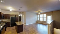 This single-floor residence features an open-concept layout that seamlessly connects the kitchen and living areas. The kitchen is outfitted with dark espresso cabinetry, a black refrigerator, and a granite-topped island that doubles as a breakfast bar, illuminated by two pendant lights hanging from the ceiling. The living area extends generously from the kitchen, highlighted by warm hardwood flooring that runs throughout the space. A ceiling fan with an integrated light fixture is centrally positioned in the living area, providing both air circulation and ambient lighting. Two large windows along the far wall allow natural light to flood the interior, contributing to the bright and open atmosphere. Neutral-toned walls complement the warm wood tones of the flooring and cabinetry. A doorway visible from the main living space suggests additional rooms beyond, such as a bedroom or bathroom. The overall design is clean and functional, blending modern finishes with a warm, inviting aesthetic suited to everyday living.