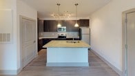 Located at 16030 Doolin Ct in Charlotte, NC, this single-floor residence offers a thoughtfully designed layout with modern finishes throughout. The kitchen serves as a standout centerpiece, featuring rich dark cabinetry with sleek bar-pull hardware, speckled granite countertops, a subway tile backsplash, and a full suite of stainless steel appliances including a refrigerator, electric range, and built-in microwave. A central island with a granite top and integrated sink is illuminated by pendant lighting, making it ideal for both meal prep and casual dining. Light wood-tone flooring extends through the kitchen and main areas, adding warmth and continuity. The living area is generously proportioned, finished with plush carpeting, and benefits from abundant natural light through a large window and a glass door that opens to an outdoor area with greenery views. A ceiling fan enhances comfort. The bedroom is carpeted as well, with a ceiling fan and a window offering views of the surrounding outdoor scenery. The bathroom is clean and functional, featuring light wood-look tile flooring, a full soaking tub with a shower surround, a toilet, and an accessible grab bar, all set against crisp white walls for a fresh, bright feel.