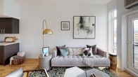 This single-floor residence showcases a thoughtfully designed layout with warm light wood flooring flowing throughout. The living area is a standout space, anchored by French doors that open to an outdoor area and frame sweeping views of the surrounding cityscape. Generous wall space accommodates curated artwork, while a wall-mounted climate control unit ensures year-round comfort. The modern kitchen is outfitted with dark cabinetry, stainless steel appliances including a French door refrigerator, electric range, dishwasher, and microwave, complemented by a stainless steel farmhouse sink, white subway tile backsplash, and open wood shelving for functional display. The bedroom offers a serene retreat, accessed through a sliding barn door, with a window framing outdoor views and ample natural light. The full bathroom features a soaking tub with shower, a dark-cabineted vanity with a white countertop, large mirror, and light wood flooring accented by a charcoal bath mat. Throughout the space, crisp white walls provide a clean, contemporary backdrop that enhances the overall bright and welcoming ambiance.