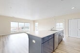 This well-appointed single-floor unit offers a thoughtfully designed open-concept layout that seamlessly connects the kitchen and living area. The kitchen is a standout feature, showcasing sleek two-tone cabinetry with slate-blue lower cabinets and crisp white upper cabinets, complemented by white quartz countertops, a full suite of stainless steel appliances including a refrigerator, range, microwave, and dishwasher, and a central island with an integrated sink. A breakfast bar with corbel supports provides additional seating and further defines the space. Light wood-look flooring runs throughout the main living areas, enhancing the bright, airy atmosphere created by large windows that frame views of the surrounding cityscape and distant mountains. In-unit stacked laundry is conveniently located within the kitchen corridor. The bedrooms are carpeted for comfort, with one featuring a generous sliding-door closet and access to an adjacent room. The bathroom is finished with wood-look flooring, a vanity with white cabinetry, a toilet, and a glass-enclosed walk-in shower. Neutral warm-toned walls throughout create a clean, contemporary aesthetic that is both welcoming and versatile.