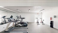 This fitness center offers a comprehensive workout environment designed to support a wide range of exercise routines. The space is laid out on a single floor with a clean, functional aesthetic, featuring white walls, a textured ceiling, and durable speckled rubber flooring throughout that provides both traction and comfort during workouts.

The cardio zone is well-stocked with multiple treadmills from True Fitness and elliptical trainers, positioned along one side of the room and reflected in a large wall-mounted mirror that enhances the sense of space. A dedicated cycling area houses several stationary spin bikes, situated near a window that allows natural light to filter in and offers a view to the outdoors.

The strength and functional training area includes a full rack of dumbbells, a colorful set of wall-mounted medicine balls, stability/exercise balls stored on a wall-mounted rack, and a flat bench. Floor mats are available for stretching or bodyweight exercises. Overhead, bright linear fluorescent lighting ensures the entire space is well-lit at all times.

Multiple entry and exit points, including a windowed door leading to an outdoor area, contribute to convenient accessibility and ventilation. The overall atmosphere is clean, organized, and purpose-built for effective fitness, making it a practical and inviting facility for all fitness levels.