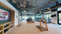 This well-appointed fitness center at 600 Saint Albans Dr in Raleigh, NC offers a generously sized, single-floor layout designed to accommodate a full range of workout disciplines. The space features an industrial-modern aesthetic, with exposed concrete columns, dark painted ceilings revealing structural ductwork, and pendant lighting that adds warmth to the environment. A charming decorative touch comes from vintage tennis rackets suspended from the ceiling, lending the space a distinctive character.

Floor-to-ceiling windows line the perimeter, flooding the interior with natural light and offering serene views of the surrounding wooded landscape. The main workout floor is finished with durable rubber and carpet-style flooring suited for both cardio and strength training.

Equipment includes a row of treadmills, elliptical machines, stair climbers, water rowing machines, and Peloton cycling bikes. A dedicated strength training area features a cable machine, dumbbell rack with bench, and rubber flooring for heavy lifting. Stall bars mounted to the walls provide additional functional training options.

A secondary zone offers built-in shelving, a flat-screen TV, and cabinetry with a beverage cooler, creating a more intimate studio or recovery area. The overall ambiance balances high-performance functionality with thoughtful design, making it an inviting and comprehensive fitness destination.