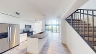 Located at 4400 West Sunrise Boulevard in Plantation, FL, this contemporary two-story loft showcases a bold, modern design defined by dramatic double-height ceilings and expansive floor-to-ceiling windows that flood the living area with abundant natural light. The main level features an open-concept layout where the kitchen flows seamlessly into the living space. The kitchen is outfitted with sleek white cabinetry, a tiled backsplash, stainless steel appliances, and a large center island with a dark stone countertop and built-in sink — ideal for both everyday cooking and entertaining. Light wood-style flooring runs throughout, complementing the crisp white walls and giving the space a clean, airy feel. A striking open-tread staircase with dark metal railings leads to the upper level, which overlooks the main floor below through an open loft-style railing. Large sliding glass doors on the main level open to a private balcony offering views of the surrounding Plantation landscape with lush palm trees. The two bathrooms are finished in a minimalist style, featuring large-format tile flooring, a frameless glass shower, a floating vanity with a stone countertop, and an oversized mirror. A generously sized walk-in closet with wire shelving provides practical storage. A hallway connects the upper-level spaces efficiently, completing this well-designed, light-filled two-floor residence.