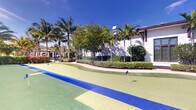 Located at 11020 Town Circle in Royal Palm Beach, Florida, this outdoor putting green offers a dedicated recreational area designed for golf enthusiasts. The space features a meticulously maintained artificial turf surface with a multi-hole putting layout, clearly delineated by vibrant color-coded turf zones in shades of green, yellow, and blue — providing both functional course variety and visual appeal.

The putting green is set against a stunning Florida backdrop, with tall, mature palm trees and lush tropical greenery framing the area and providing natural shade. A manicured hedge line and ornamental shrubs create a clean boundary between the green and the adjacent paved pathway, enhancing the overall aesthetic.

The neighboring structure — a white stucco building with a dark hip roof, rich wood-toned window trim, and large shuttered windows — adds an elegant architectural presence to the setting. Under the expansive South Florida sky, the space is bathed in abundant natural sunlight, giving it a bright, resort-like atmosphere. The combination of professional-grade turf, tropical landscaping, and refined architectural surroundings makes this putting green a standout recreational amenity well-suited for practice or leisure.