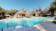 This community amenity space centers around a generously sized, freeform resort-style swimming pool with a smooth concrete and stone-tile surround. The pool deck is outfitted with lounge chairs, shaded umbrella seating, and lush tropical plantings — including striking sago palms — that create natural focal points throughout the outdoor area. A second, smaller pool is also visible, offering additional recreational options for residents.

The clubhouse building anchors the space with a charming architectural design that combines natural stone masonry, warm wood siding, a pitched shingle roof, and a prominent chimney. Large French-style windows and a covered veranda along the front face of the building invite natural light and provide a welcoming transition between the indoor and outdoor environments. Manicured landscaping with ornamental beds and mature trees frames the entrance and driveway approach.

Adjacent to the pool, a substantial wood pergola shelters an outdoor kitchen station equipped with multiple built-in grills set into a stone surround — ideal for communal gatherings and al fresco dining. The entire complex is surrounded by mature trees that offer shade and privacy, while the open sky above ensures an abundance of natural light throughout the day. The cohesive design and well-maintained grounds reflect a thoughtfully planned community amenity environment.