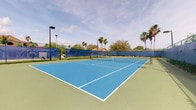 This outdoor tennis facility showcases a full-size hard court finished in a striking two-tone blue and green surface, with crisp white lines clearly demarcating the playing areas. The court is equipped with a standard net positioned at regulation height, ensuring readiness for competitive or recreational play. Surrounding the court is a chain-link perimeter fence fitted with blue windscreen panels, which provide wind protection and help keep balls within the playing area during matches. Overhead lighting fixtures mounted on tall poles indicate the court is designed to support evening play, extending usability beyond daylight hours. The facility is framed by mature palm trees and dense tropical vegetation, creating a pleasant and shaded perimeter that adds to the outdoor ambiance. Neighboring residential structures are visible beyond the fence line, suggesting the court is part of a community or residential amenity. The open-air setting, combined with the well-kept playing surface and surrounding greenery, gives the facility a welcoming and functional character suited to regular use by players of varying skill levels.