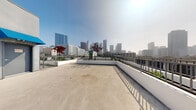 Located in the heart of Los Angeles at 744 Hartford Ave, this rooftop terrace offers a commanding presence above the city with sweeping, unobstructed views of the Downtown Los Angeles skyline. The open-air deck is finished in smooth concrete and wraps around a central rooftop mechanical structure, creating a generous and flexible outdoor space suited for a variety of uses.

A sleek iron railing lines the perimeter, providing safety while preserving the dramatic cityscape sightlines in every direction. Raised white-painted planters add touches of greenery, featuring ornamental trees and drought-tolerant plants that bring life to the urban setting. The layout of the terrace allows for multiple vantage points, from close-up views of neighboring mid-rise buildings to distant panoramas of Downtown LA's iconic high-rise towers.

The rooftop structure itself is finished in a clean grey stucco exterior with bold blue canopies accenting doorway entries, providing both visual interest and functional shade. Access is provided via clearly marked entry doors, with elevator access also available. The terrace's open, uncluttered design makes it highly adaptable, offering significant potential for outdoor gatherings, events, or amenity use. The abundance of natural light and the ever-present Los Angeles skyline backdrop make this rooftop a truly distinctive urban outdoor space.