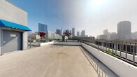 Located in the heart of Los Angeles at 744 Hartford Ave, this rooftop terrace offers a commanding presence above the city with sweeping, unobstructed views of the Downtown Los Angeles skyline. The open-air deck is finished in smooth concrete and wraps around a central rooftop mechanical structure, creating a generous and flexible outdoor space suited for a variety of uses.

A sleek iron railing lines the perimeter, providing safety while preserving the dramatic cityscape sightlines in every direction. Raised white-painted planters add touches of greenery, featuring ornamental trees and drought-tolerant plants that bring life to the urban setting. The layout of the terrace allows for multiple vantage points, from close-up views of neighboring mid-rise buildings to distant panoramas of Downtown LA's iconic high-rise towers.

The rooftop structure itself is finished in a clean grey stucco exterior with bold blue canopies accenting doorway entries, providing both visual interest and functional shade. Access is provided via clearly marked entry doors, with elevator access also available. The terrace's open, uncluttered design makes it highly adaptable, offering significant potential for outdoor gatherings, events, or amenity use. The abundance of natural light and the ever-present Los Angeles skyline backdrop make this rooftop a truly distinctive urban outdoor space.