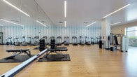 This fitness center is a thoughtfully designed workout space that combines functionality with a clean, modern aesthetic. The room features light hardwood flooring throughout, providing a warm, polished foundation that complements the bright, white ceiling overhead. Linear LED light fixtures run parallel across the ceiling, delivering consistent, even illumination throughout the space.

A full-length mirror spans the length of one wall, visually expanding the room and allowing users to monitor their form during exercise. The opposite wall serves as a striking design focal point, featuring a nature-inspired wallcovering depicting a birch tree forest in soft blue and white tones, adding visual depth and a calming ambiance to the environment.

The cardio section is well-stocked with a row of treadmills and elliptical machines, all equipped with individual digital display consoles. The free weight area includes a rack of dumbbells and a flat weight bench positioned on a protective rubber mat, accommodating a variety of strength training exercises. A standing workstation or display unit is positioned near the far wall, and a waste receptacle is conveniently located nearby. The overall layout is open and spacious, allowing for comfortable movement between equipment.