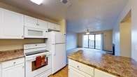 This single-floor residence offers a practical and comfortable layout with an open-concept design that seamlessly connects the kitchen and living area. The kitchen is well-equipped, featuring white cabinetry, a full-size refrigerator, an electric range, and an over-the-range microwave. A peninsula with a granite-style countertop provides additional workspace and serves as a natural divider between the kitchen and the adjoining living space. The flooring transitions from warm hardwood-style in the kitchen to plush carpeting in the living area, adding texture and comfort underfoot. The living area is anchored by a ceiling fan and benefits from abundant natural light streaming through a large sliding glass door, which opens directly to an outdoor balcony. This seamless indoor-outdoor connection enhances the sense of openness throughout the space. The textured ceilings add a classic architectural detail, while neutral wall tones throughout create a versatile backdrop suited to a variety of interior styles. Overall, the space strikes a balance between functionality and livability, making it an inviting and well-appointed residence.