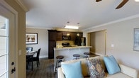 Located on State Street, this single-floor residence offers a thoughtfully designed open-concept layout that seamlessly connects the kitchen, dining, and living areas. The kitchen is a standout feature, boasting rich dark cabinetry that extends to the upper walls, black appliances including a range and built-in microwave, and a light-toned granite breakfast bar with industrial-style bar stools — ideal for casual dining or entertaining. Pendant lighting hangs above the bar, complementing the overhead fixtures and adding a warm, inviting ambiance. Crown molding frames the smooth white ceilings throughout, lending an elegant, finished quality to the interior. The living area flows naturally from the kitchen, anchored by a comfortable sofa with decorative accent pillows in soft blues and earth tones. A ceiling fan is visible overhead, contributing to practical airflow and comfort. The dining space, positioned to one side, accommodates a full table and chair set. Neutral gray walls and dark hardwood-style flooring run throughout, creating a cohesive, modern aesthetic. A hallway visible in the background suggests additional private rooms beyond the main living space.