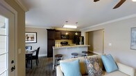 Located on State Street, this single-floor residence offers a thoughtfully designed open-concept layout that seamlessly connects the kitchen, dining, and living areas. The kitchen is a standout feature, boasting rich dark cabinetry that extends to the upper walls, black appliances including a range and built-in microwave, and a light-toned granite breakfast bar with industrial-style bar stools — ideal for casual dining or entertaining. Pendant lighting hangs above the bar, complementing the overhead fixtures and adding a warm, inviting ambiance. Crown molding frames the smooth white ceilings throughout, lending an elegant, finished quality to the interior. The living area flows naturally from the kitchen, anchored by a comfortable sofa with decorative accent pillows in soft blues and earth tones. A ceiling fan is visible overhead, contributing to practical airflow and comfort. The dining space, positioned to one side, accommodates a full table and chair set. Neutral gray walls and dark hardwood-style flooring run throughout, creating a cohesive, modern aesthetic. A hallway visible in the background suggests additional private rooms beyond the main living space.