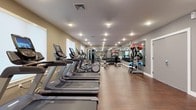 This fitness center is designed to provide a comprehensive workout environment within a single, thoughtfully organized space. The room features a row of modern cardio machines — including treadmills and ellipticals — positioned along one side, each equipped with individual display screens for an enhanced exercise experience. Recessed and flush-mounted ceiling lights illuminate the space evenly, creating a clean, energizing atmosphere throughout.

The opposite wall is lined with large mirrors that visually expand the room and allow users to monitor their form during exercise. A dedicated free weights section includes a full rack of dumbbells and an assortment of medicine balls and stability balls neatly arranged on standing racks, supporting a wide range of strength and functional training routines. Additional cable and resistance machines are visible toward the rear of the space, offering further versatility.

The warm hardwood-style flooring contrasts with the neutral, light gray walls, contributing to a welcoming and professional ambiance. Natural light filters in through windows along one wall, complementing the overhead lighting. The layout is open and organized, promoting ease of movement between equipment zones. Overall, this fitness center is well-appointed and suitable for a variety of workout routines.