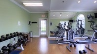 This fitness center offers a clean, functional environment designed to support a variety of workout routines. The space features warm hardwood flooring throughout, complemented by soft sage green walls and crisp white crown molding that lend a polished, welcoming feel. Large floor-to-ceiling mirrors line one wall, visually expanding the room and allowing users to monitor their form during exercise. The ceiling is finished with recessed lighting alongside a central flush-mount fixture, ensuring the space is well-lit at all times. Cardio equipment, including elliptical machines and additional aerobic trainers, is neatly arranged along the mirrored wall, with a mounted flat-screen television nearby for entertainment during workouts. A dedicated free weights area with a rolling dumbbell rack provides options for strength training. A small waste bin and accessory station are conveniently positioned near the entrance. Natural light filters in through a windowed door that leads to an outdoor area, adding an airy quality to the interior. The overall layout is thoughtfully organized, maximizing the available floor space to accommodate multiple users simultaneously while maintaining an open, uncluttered atmosphere.