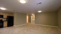 This single-floor space is characterized by a spacious, open main living area finished with neutral beige walls and white trim baseboards that create a clean, understated aesthetic. The flooring consists of light-toned tile or vinyl, contributing to a bright and easy-to-maintain environment. Flush-mount ceiling light fixtures illuminate the space evenly, providing consistent artificial lighting throughout. A ceiling-mounted HVAC vent ensures climate control is integrated directly into the layout.

Toward the rear of the main area, two doorways provide access to additional spaces. One opening leads to what appears to be a well-lit side room or hallway, while the other reveals an in-unit laundry area, visibly equipped with a washer and dryer side by side — a practical and convenient feature. The walls throughout are smooth and uniformly painted, lending a move-in-ready feel to the interior.

The overall layout is open and functional, with no partition walls dividing the main living zone, allowing for flexible furniture arrangement. The space maintains a straightforward, residential character suited to everyday living needs.