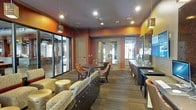 This single-story space serves as a well-appointed communal lounge and activity area. The interior is defined by its rich, warm color palette, with deep reddish-orange and amber-toned accent walls that create an inviting and vibrant atmosphere throughout. Dark wood-toned flooring runs the length of the space, complementing the earthy tones of the walls and furniture.

The lounge area is thoughtfully furnished with a variety of seating options, including a generously sized upholstered sofa adorned with floral patterned cushions, paired with rounded metallic accent ottomans. A separate seating cluster features a dark circular table surrounded by patterned armchairs, ideal for small group gatherings or casual meetings.

Along one side of the space, a dedicated workstation area is equipped with computer terminals and a counter surface, functioning as a business center. Large glass panel walls on one side allow for visibility into an adjacent room, contributing to an open and connected feel. Recessed lighting and pendant fixtures illuminate the space evenly, while multiple doorways provide access to additional rooms. The overall design balances functionality with a warm, welcoming ambiance.