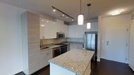 This single-floor kitchen showcases a thoughtfully designed, contemporary layout that balances style and functionality. The space is anchored by a generously sized central island topped with speckled granite countertops, offering ample workspace and a natural gathering point. Matching granite surfaces extend throughout the kitchen, providing a cohesive and polished aesthetic.

Cabinetry is a standout feature, combining crisp white upper cabinets with light wood-toned lower cabinets that add warmth and visual contrast. The clean-lined, handle-accented doors contribute to the modern sensibility of the space. Stainless steel appliances, including a refrigerator, range with oven, microwave, and dishwasher, are seamlessly integrated throughout.

Overhead, a combination of adjustable track lighting and elegant pendant lights suspended above the island ensures the space is well-lit and visually dynamic. Dark hardwood flooring grounds the lighter tones of the cabinetry and counters, creating a striking contrast. A neutral backsplash completes the refined look, making this kitchen a highly functional and visually appealing space.