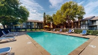 This outdoor amenity space centers around a generously sized swimming pool with clear turquoise water, framed by a wide concrete and paved sun deck. Blue lounge chairs are arranged along the pool's perimeter, offering ample space for sunbathing and relaxation, while yellow patio umbrellas provide convenient shaded seating areas for those seeking relief from the sun. The pool is fully enclosed by a black iron security fence, ensuring a safe and well-defined recreational environment. A brick-constructed covered pavilion sits adjacent to the pool area, featuring a vaulted, beam-accented ceiling and an open-air design that allows for natural ventilation. The pavilion houses a round picnic table, making it a practical gathering spot for outdoor dining and socializing. The surrounding landscape is enriched by a collection of large, mature trees that cast natural shade over portions of the deck and create a lush, park-like atmosphere. The multi-story residential buildings visible in the background feature balconies and a classic brick exterior, contributing to the cohesive architectural character of the community. Together, these elements combine to create a vibrant and functional outdoor recreational space well-suited for both leisurely relaxation and social gatherings.