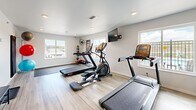 Located at 7175 Dunsmore Rd in Rapid City, South Dakota, this fitness room is a compact yet thoroughly equipped exercise space designed to support a well-rounded workout routine. The room features light wood-look flooring throughout, crisp white walls, and recessed lighting that creates a clean, energizing atmosphere. Multiple windows along the walls allow generous natural light to fill the space while offering views of the surrounding area.

The cardio zone is anchored by treadmills and an elliptical trainer, all positioned on protective floor mats. A wall-mounted flat-screen television provides entertainment during workouts. A full-length wall mirror visually expands the room and assists with form and technique. The free weights area includes a well-stocked dumbbell rack with a complete range of weights, complemented by wall-mounted motivational signage.

For functional and flexibility training, the space features a wall-mounted storage rack holding medicine balls and stability balls in multiple sizes, along with a hanging workout mat. Industrial-style wall fans ensure proper air circulation throughout the room. A seated bench and additional floor mats round out the functional training options. The room is accessed through a solid-framed glass-inset door, contributing to the space's modern, purposeful aesthetic.