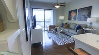 This well-appointed single-floor residence offers a thoughtfully designed living area that balances comfort and style. Rich, dark hardwood flooring extends throughout the main living space, providing a warm and grounding foundation for the interior. A ceiling fan is centrally positioned overhead, adding both a functional and decorative element to the room.

The living area is anchored by a plush gray sofa accented with navy and gold throw pillows, complemented by a pair of leather accent chairs and a sleek glass-top coffee table. A large flat-screen television is mounted on the wall beside a clean-lined white media console, creating a well-organized entertainment zone. Decorative artwork, including a striking bird portrait in warm tones, adds a touch of personality to the walls.

Natural light floods the interior through a full-height sliding glass door that opens onto a private balcony, offering open outdoor views that extend to the horizon. The balcony enhances the airy, expansive feel of the space. Neutral sage-green walls provide a calm, neutral backdrop that ties together the contemporary furnishings. A patterned blue-and-white area rug defines the seating zone and adds visual interest, completing the cohesive and inviting atmosphere of this residence.