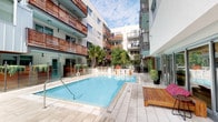 This striking contemporary courtyard space is centered around a pristine resort-style swimming pool, offering an elevated outdoor living experience. The pool area is framed by a multi-story building with clean, modern architectural lines, featuring a combination of white exterior panels and warm wood-toned horizontal balcony railings that add texture and visual depth to the façade. Private balconies with cable railings overlook the courtyard, creating a sense of community while maintaining individual privacy.

The pool deck is finished in light-toned stone pavers, providing an elegant and cohesive surface throughout the outdoor space. A standout feature is the lush vertical green wall, which introduces a vibrant natural element against the modern built environment. Tall tropical palms and potted plants are strategically placed throughout the courtyard, enhancing the resort-like atmosphere.

Outdoor seating areas are thoughtfully arranged around the pool, including a large stone-topped bar or counter with contemporary seating, as well as a dining table with white chairs. Teak-style sun loungers offer additional relaxation spots. Large sliding glass doors connect the interior spaces to the outdoor courtyard, seamlessly blending indoor and outdoor living. String lights hint at the space's appeal during evening hours, making it equally inviting day and night.