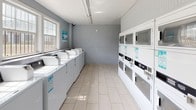 Located at 3025 E Park Row Dr in Arlington, Texas, this single-floor community laundry facility is designed to accommodate a high volume of laundry needs efficiently. The space is laid out in a long, open rectangular floor plan with light-colored ceramic tile flooring throughout, contributing to a clean and practical environment.

Along one wall, a generous row of top-loading washing machines is neatly arranged, providing ample capacity for simultaneous use. Opposite these, stacked commercial dryer units line the adjacent wall, maximizing the use of vertical space and offering multiple drying options. A separate section of the facility houses additional front-loading washers, expanding the overall capacity further.

Natural light filters into the space through several windows fitted with blinds, brightening the interior and creating a more welcoming atmosphere. The walls are painted in a calm gray tone, and overhead fluorescent lighting ensures consistent illumination throughout all hours of use. Informational signage is posted at key points to guide users through machine operation.

A fire extinguisher is visibly mounted, reflecting attention to safety standards. The facility also includes a countertop area, providing a convenient surface for folding or sorting laundry. Overall, this laundry facility is a well-organized, functional space built to serve the practical needs of its community effectively.