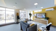 This single-floor lounge is designed with a contemporary aesthetic, combining vibrant yellow accent walls with a clean, neutral palette of whites and grays. Large floor-to-ceiling windows line one side of the room, flooding the space with abundant natural light and offering open views of the surrounding urban environment. Recessed panel lighting in the white ceiling further enhances the bright, welcoming atmosphere.

The seating area is anchored by a pair of sleek gray sofas accented with mustard yellow throw pillows, arranged around a low white coffee table to create an inviting, social environment. A cushioned accent chair adds additional seating flexibility. Across from the lounge area, a large flat-screen television is mounted above a warm wood-toned media console, framed by the bold yellow feature wall and complemented by framed artwork.

Lush potted indoor trees are positioned throughout the space, introducing a natural element that softens the modern décor. The light gray carpeting ties the room together, providing a cohesive and comfortable foundation. Overall, this lounge offers a polished, energetic environment well-suited for both relaxation and casual gatherings.