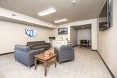 This single-floor communal lounge is designed with a clean, modern aesthetic, featuring neutral gray walls, light carpeted flooring, and recessed panel lighting that creates a bright, welcoming atmosphere. The main lounge area is generously sized, accommodating multiple seating arrangements including gray tufted sofas and upholstered armchairs grouped around low wooden coffee tables. Board games are available for resident use, adding a casual, social element to the space. Decorative wall art in blue and coastal tones adds warmth to the otherwise understated interior. A wall-mounted flat-screen television serves as a central focal point, viewable from multiple seating areas. A window along one wall allows natural light to filter in, offering views of the surrounding area. The built-in kitchenette features white shaker-style cabinetry, a light quartz countertop, a stainless steel sink, a microwave, and a compact refrigerator, providing convenient amenities for occupants. An adjacent alcove opens into a game room equipped with a foosball table, extending the recreational offerings of the space. A commercial-grade door with a narrow vertical window provides secure access to the lounge, reflecting the space's communal facility design.