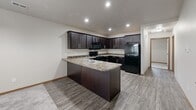 This single-floor, 2-bedroom, 1-bathroom home offers a practical and comfortable layout designed for everyday living. The open-concept living and dining area is finished with soft carpet flooring and features a ceiling fan, a decorative chandelier, and a textured ceiling. Natural light filters in through multiple windows, offering views of the surrounding greenery. The space flows seamlessly into a well-equipped kitchen, which boasts rich dark espresso cabinetry, granite-style countertops, a peninsula with a double sink, and a full suite of black appliances including a range, microwave, and refrigerator. Recessed lighting and wood-look tile flooring complete the kitchen's polished look. Both bedrooms are carpeted and include ceiling fans, with the primary bedroom offering a generous footprint. Each bedroom includes closet space for ample storage. The full bathroom features a combined tub and shower, a dark-cabinet vanity, and wood-look flooring in a clean, neutral palette. A dedicated laundry room with side-by-side washer and dryer units and overhead wire shelving adds everyday convenience. The home's cohesive neutral tones and functional design make it a comfortable and move-in-ready living space.