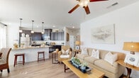 This single-story home offers a thoughtfully designed open-concept layout that seamlessly connects the living, dining, and kitchen areas. The living room features vaulted ceilings with a ceiling fan, light wood-look flooring, and large windows that invite natural light. The kitchen is well-equipped with rich dark cabinetry extending to the ceiling, granite countertops, stainless steel appliances, and a breakfast bar with seating. Adjacent to the kitchen, the dining area accommodates a round table beneath an elegant chandelier, with windows dressed in sheer white curtains providing a bright, airy ambiance. The bedrooms offer comfortable retreats; one features plush carpet, a ceiling fan, and a window with natural light, while another presents a coastal-inspired aesthetic with light walls and decorative accents. The bathroom includes dark cabinetry with granite countertops, a large mirror, and a tub-shower combination with tile surround. A dedicated laundry room houses a full-size washer and dryer with additional cabinetry for storage. Throughout the home, light wood-look flooring, white walls, and consistent finishes create a cohesive, warm, and inviting atmosphere.