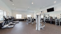 This fitness center offers a spacious, single-floor layout designed to accommodate a wide variety of workout needs. The interior is bright and clean, with white walls and recessed lighting that create a well-lit, motivating environment. Large windows along the walls allow natural light to fill the space, contributing to an open and energizing atmosphere.

The flooring consists of durable, slip-resistant rubberized material in a speckled pattern, ideal for high-traffic exercise areas and providing stability during workouts. The equipment selection is extensive and modern, featuring a row of treadmills for cardiovascular training alongside a variety of cable and selectorized strength machines suited for targeting multiple muscle groups. A dedicated free weights area with a mirror wall rounds out the strength-training options, allowing users to monitor their form.

The layout is thoughtfully arranged to provide ample space between machines, ensuring ease of movement and a comfortable workout experience. A wall-mounted television is also present, adding to the amenity. The overall design is functional and professional, making this fitness center a practical and fully equipped resource for daily exercise.