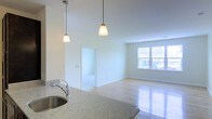 This single-floor space showcases an open-concept layout that seamlessly connects the kitchen and living areas. The kitchen features speckled granite countertops with an undermount stainless steel sink and a classic arc faucet. Two pendant lights with frosted glass shades hang overhead, providing focused task lighting while adding a decorative touch. The light hardwood flooring extends throughout, contributing to a clean, cohesive aesthetic. The adjacent living area is bright and airy, benefiting from a large window that allows ample natural light to fill the room and offers views of the surrounding greenery. Crisp white walls and white trim throughout the space enhance the sense of openness. A doorway visible from the main living area leads to an additional room, suggesting a flexible floor plan. The overall ambiance is fresh, neutral, and move-in ready, offering a versatile canvas suitable for a variety of furnishing and décor styles.