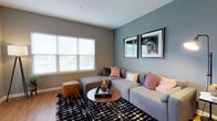 This single-floor residence showcases a thoughtfully designed interior with a cohesive modern aesthetic. The living room is bright and welcoming, featuring large windows that flood the space with natural light, warm hardwood-style flooring, and cool-toned accent walls that add depth and character. A spacious sectional sofa anchors the room, complemented by a round coffee table and bold patterned area rug. An entry area adjacent to the living room offers additional open space with a private door and stylish sideboard console. The dining area flows seamlessly from the living room and features a round wooden dining table with leather chairs beneath a brushed nickel chandelier, creating an inviting atmosphere for meals and gatherings. The kitchen is equipped with rich dark wood cabinetry, granite-style countertops, and a full suite of stainless steel appliances, including a gas range, microwave, and refrigerator, all set over the same warm hardwood-style floors. The bedroom is a serene retreat highlighted by a bold dark blue accent wall, plush carpeting, ample natural light from a large window, and convenient access to the bathroom. The bathroom features light tile flooring, dark wood vanity cabinetry, a wide mirror with overhead lighting, and both a soaking tub and a separate shower.