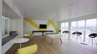 This single-floor common space showcases a contemporary, energetic design anchored by striking bold yellow geometric stripes painted across a white accent wall. A wall-mounted flat-screen television is centered on this feature wall, while a shuffleboard table positioned beneath it adds a recreational focal point to the room. The space is thoughtfully arranged with a mix of seating options, including bright yellow lounge chairs paired with round white tables, creating casual conversation areas. A white ping-pong table with natural wood legs serves as a central gathering point on the light hardwood flooring that runs throughout. Along the perimeter, floor-to-ceiling windows flood the interior with abundant natural light while framing panoramic outdoor views of the surrounding landscape. Dark modern chairs and tall bistro-style tables are positioned near the windows, offering a more relaxed vantage point to enjoy the scenery. Recessed ceiling lighting complements the natural light, ensuring the space remains bright and inviting. Overall, the room exudes a lively, social atmosphere well-suited for recreation and casual gatherings.