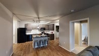 This single-story space showcases an open-concept kitchen designed for both functionality and style. The kitchen is equipped with dark espresso-toned cabinetry that extends along two walls, offering generous storage. Contrasting light countertops provide a clean, modern aesthetic, while black appliances — including a refrigerator, range, and built-in microwave — add a cohesive, contemporary finish. A central kitchen island serves as a focal point, accommodating seating for multiple guests with metal bar stools, making it ideal for casual dining or entertaining. Overhead, a modern globe pendant light fixture adds a decorative touch above the island, complementing the track lighting positioned nearby. The warm hardwood-style flooring extends throughout the main living area, adding texture and warmth to the space. A doorway visible from the kitchen leads to what appears to be a dedicated laundry area with a washer and dryer. An additional doorway hints at further rooms beyond, suggesting a well-connected layout. The neutral gray wall tones throughout create a versatile, move-in-ready atmosphere.