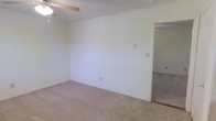 This single-floor residential space offers a straightforward and functional layout designed for comfortable everyday living. The interior is finished with soft, neutral-toned wall paint that creates a bright and airy atmosphere throughout. The main living area features wall-to-wall carpeting in a light beige tone, providing a warm and cushioned underfoot experience. A ceiling fan is mounted overhead, offering both lighting and air circulation to keep the space comfortable year-round. A wall-mounted thermostat is conveniently located for easy climate control. The room flows seamlessly into an adjacent space through an open doorway, suggesting a connected and practical floor plan. Electrical outlets are positioned at standard intervals along the walls, offering versatile placement options for furniture and appliances. The clean, unadorned walls provide a blank canvas, making the space highly adaptable to a variety of interior design preferences. The overall ambiance is simple, clean, and move-in ready, appealing to those who appreciate a minimalist foundation they can personalize to their own taste.