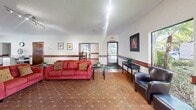 Located at Heather Terrace in Fremont, CA, the West Downstairs Club House is a generously sized single-floor community facility designed to serve as a welcoming gathering space for residents. The interior is defined by a cohesive design aesthetic, featuring terracotta tile flooring in the entryway and transitional areas, warm patterned carpeting throughout the main lounge and dining spaces, and a consistent red tile wainscoting that runs along the walls, adding a distinctive decorative touch.

The entry foyer welcomes visitors with large windows overlooking lush greenery and a classic coat rack, setting a comfortable tone. The spacious lounge area is furnished with seating including a red sofa and leather armchairs, a wall-mounted flat-screen TV, and large windows that flood the space with natural light and views of the surrounding landscaping. Track lighting throughout provides ample illumination.

Multiple dining areas accommodate gatherings of various sizes, with both a large dark-wood table and a smaller wood dining set available. A brick fireplace serves as a focal point in the main dining room, which also features sliding glass door access to an outdoor pool area. A kitchenette with appliances supports event hosting. The textured ceilings and framed artwork throughout complete the inviting, community-oriented ambiance.