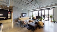 This single-floor industrial-modern loft showcases a striking open-plan layout defined by raw exposed concrete walls, polished concrete floors, and soaring ceilings with fully exposed ductwork and pendant lighting. The main living area is anchored by a generous sectional sofa arrangement around a gold drum coffee table atop a bold geometric rug, with a wall-mounted flat-screen TV and a sleek wood media console against the concrete feature wall. Floor-to-ceiling steel-framed windows and glass doors flood the space with abundant natural light and offer expansive urban skyline views, with direct access to an outdoor terrace. A dedicated lounge nook features a dramatic floor-to-ceiling wall of vinyl records housed in custom shelving, complemented by a warm cognac leather sofa and lush potted plants. A separate zone is accessed through elegant black steel-framed glass partition doors, revealing warm hardwood flooring, a large conference or dining table, and a connected kitchen area. The entryway features a structural concrete column alongside a glass-paneled door leading to the terrace. Throughout, the space blends industrial rawness with carefully curated furnishings, greenery, and warm accent tones to create a sophisticated yet welcoming ambiance.