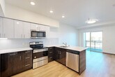 Located at 3118 6th Avenue in Tacoma, WA, this modern one-bedroom, one-bathroom unit offers a thoughtfully designed layout across a single floor. Light-toned hardwood-style flooring runs throughout, creating a cohesive and clean aesthetic from room to room.

The open-concept kitchen is a standout feature, showcasing a two-tone design with dark wood lower cabinetry and crisp white upper cabinets. Stainless steel appliances — including a range, over-the-range microwave, dishwasher, and refrigerator — are complemented by white quartz countertops and a subway tile backsplash. A peninsula with matching dark cabinetry provides additional counter space and separates the kitchen from the living area. Recessed lighting illuminates the space, while a sliding glass door leads to a private balcony, bringing in natural light and outdoor views.

The spacious primary bedroom offers a generous footprint, neutral white walls, and a sliding-door closet with built-in shelving and hanging rods, providing ample storage.

The full bathroom is clean and functional, featuring a soaking tub with shower, a dark wood vanity with white countertop, and the same light wood-style flooring found throughout the unit. A hallway connects the rooms, contributing to the efficient and well-organized flow of the space.