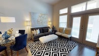 This single-story space showcases a thoughtfully designed living area that balances contemporary style with comfortable functionality. The room features notably high ceilings, enhanced by tall windows that flood the space with generous natural light, creating an open and airy atmosphere throughout.

The living area is anchored by a bold black-and-white geometric diamond-patterned area rug, which grounds the space and adds graphic contrast. A charcoal gray sofa, adorned with an assortment of decorative pillows in gold, cream, and teal tones, serves as the central seating element. A white faux-fur bench positioned in front of the sofa adds a textural focal point. Two neutral-toned accent chairs with gold throw pillows flank the window side, providing additional seating while taking advantage of the natural light.

A large abstract canvas in silver and gold tones hangs prominently on the main wall, complemented by matching perforated floor lamps on either side that add warm ambient lighting. The flooring is light wood, contributing to the overall bright and modern aesthetic. The overall ambiance is polished, welcoming, and well-balanced in both color and form.