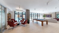 Located at 427 S Hill St in Downtown Los Angeles, this single-floor clubroom is a well-appointed amenity space designed for both socializing and relaxation. The main gathering area is anchored by light hardwood flooring and floor-to-ceiling glass doors that open to an outdoor terrace, flooding the interior with natural light and providing views of the surrounding courtyard. A wood-framed billiards table serves as a central focal point, complemented by a seating arrangement of bold red accent chairs grouped around a low coffee table beneath a sculptural circular pendant light.

Adjacent to the main area, a dedicated lounge features a large wall-mounted flat-screen TV, a plush sectional sofa, accent chairs, and a snack display cabinet, creating an inviting media retreat. A wet bar area offers dark wood cabinetry with granite countertops, open shelving, and pendant lighting, providing an ideal space for entertaining. A compact private meeting room with a round dark table, coordinating chairs, and carpeted flooring offers a quieter setting for small gatherings. Throughout, contemporary furnishings, greenery, and a neutral yet warm color palette contribute to a cohesive, upscale ambiance.