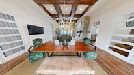 Located in Northport, Alabama, this thoughtfully designed single-floor space offers a generous, open layout that seamlessly connects its key areas. The centerpiece of the space is a striking living room crowned by a richly stained wood coffered ceiling with recessed lighting, lending an air of warmth and architectural distinction. A dramatic floor-to-ceiling stacked stone fireplace with a rustic wood mantel anchors the room, complementing the rich hardwood flooring found throughout. Large French doors and abundant windows flood the interior with natural light while providing views to the outdoors. The dining area is highlighted by a statement antler chandelier suspended above a warm wood table surrounded by patterned upholstered chairs, creating an inviting gathering space. Adjacent to the dining area, the kitchen features white shaker cabinetry, stainless steel appliances, a spacious white quartz bar with seating, and striking brass drum pendant lights that add a touch of elegance. A secondary living area offers wall-mounted televisions, a console table, and additional seating, providing a relaxed, functional retreat. The consistent use of hardwood floors, neutral walls, and curated décor throughout creates a cohesive, welcoming ambiance suited for community gatherings and everyday use.